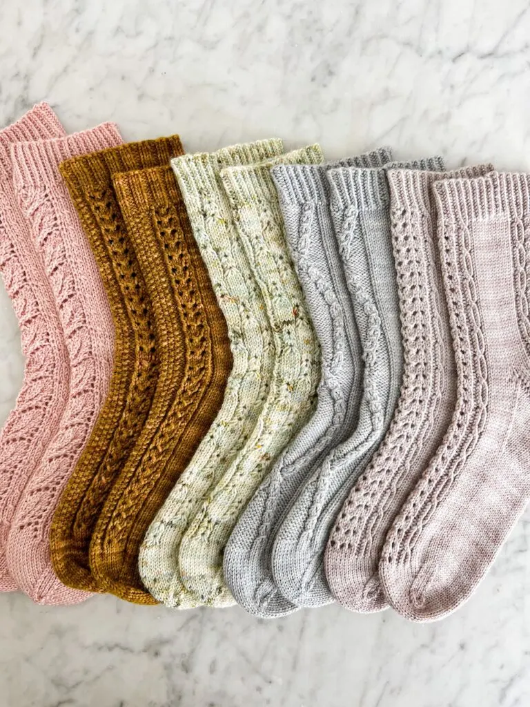 A top-down image of five pairs of socks forming a rainbow in pink, caramel, mint, ice blue, and lavender. The socks all have different textures and their toes point to the left.