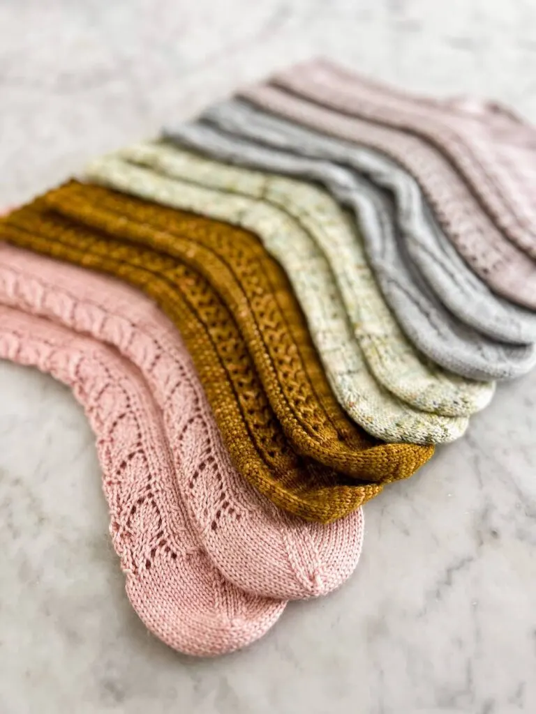A rainbow of handknit socks is laid out on a marble countertop with all the toes pointing to the left. This image is shot from the pink toes on the left-most side of the rainbow and angled back along all the socks.