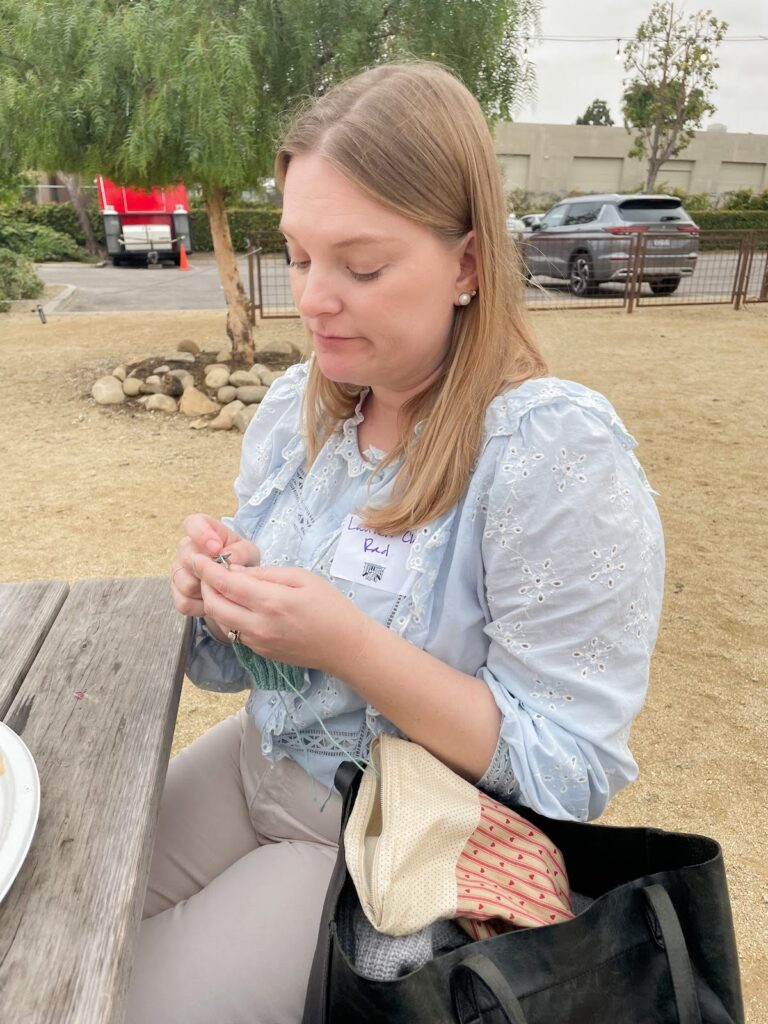 A blonde white woman in a blue ruffled shirt (that's me!) scowls at her knitting with an exasperated expression.