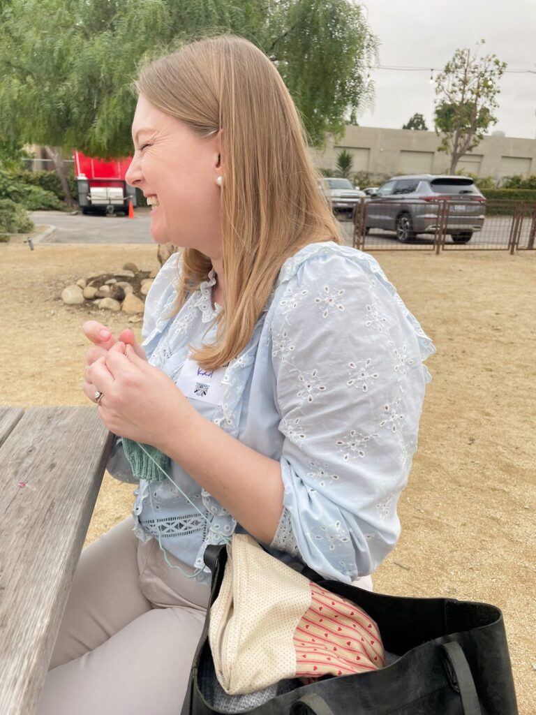 A blonde white woman in a blue ruffled shirt (that's me!) cracks up with laughter while trying to knit a sock.