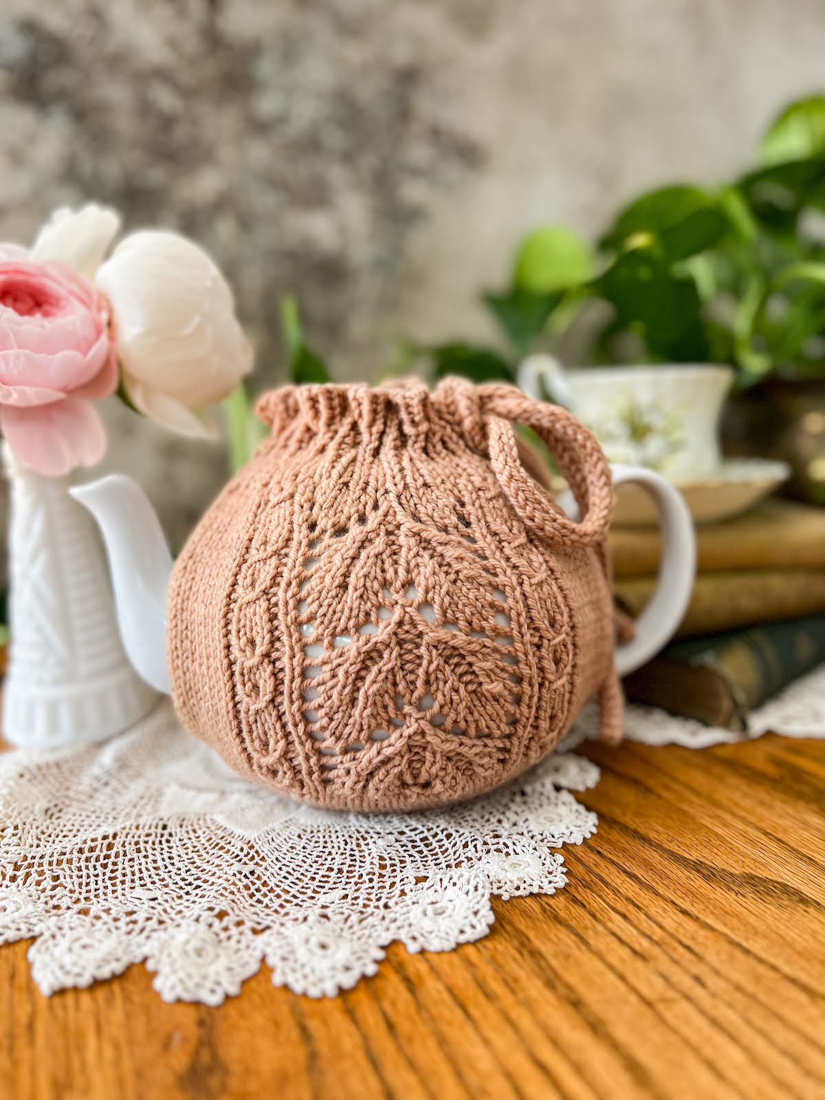 A straight-on view of the side of a clay-colored handknit tea cozy tied over a white teapot. The Emily's Garden Tea Cozy features panels of leafy lace and coin lace on each side of the cozy, and it's held together with a drawstring at the top.