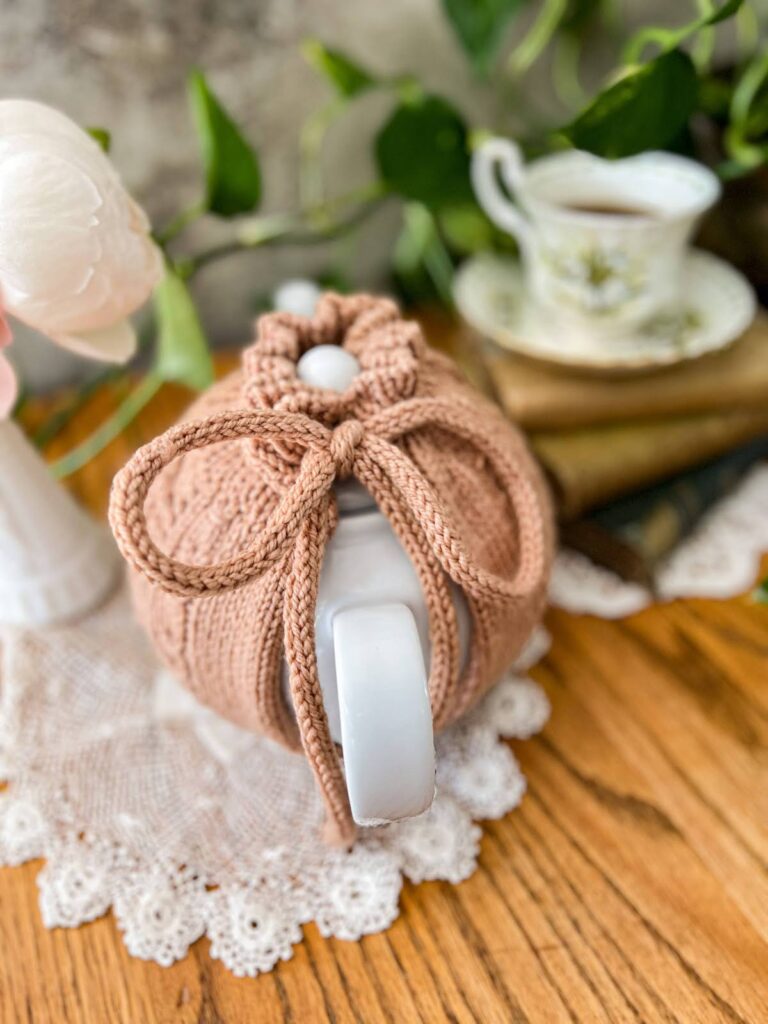 A top-down image of a clay-colored tea cozy tied onto a white teapot. The Emily's Garden Tea Cozy is held together with an i-cord drawstring, which is the focus of this photo.