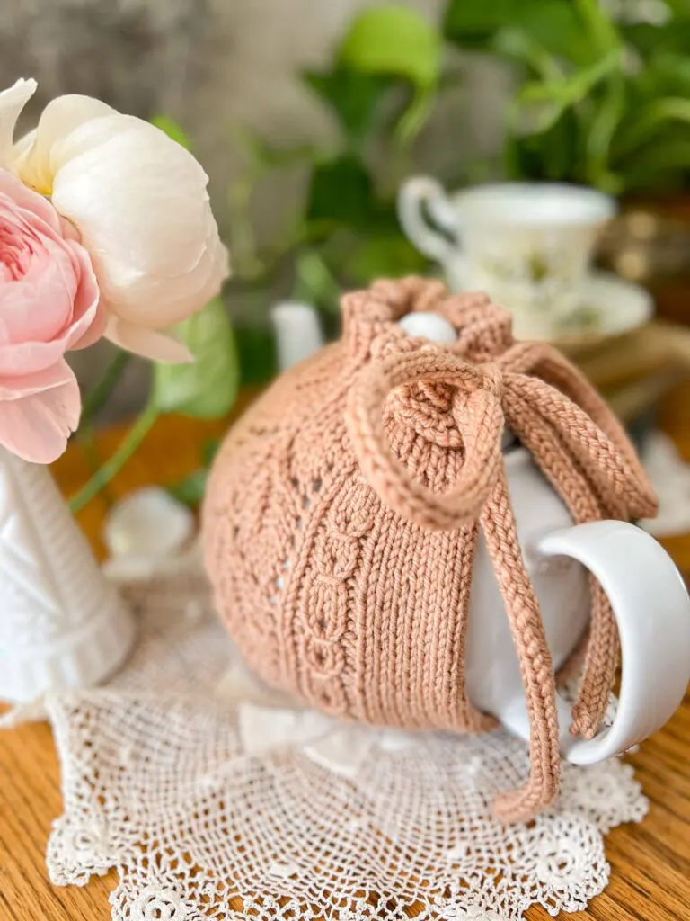 An angled image of a clay-colored tea cozy on a white teapot, shot from the handle toward the spout (which is blurred in the background).