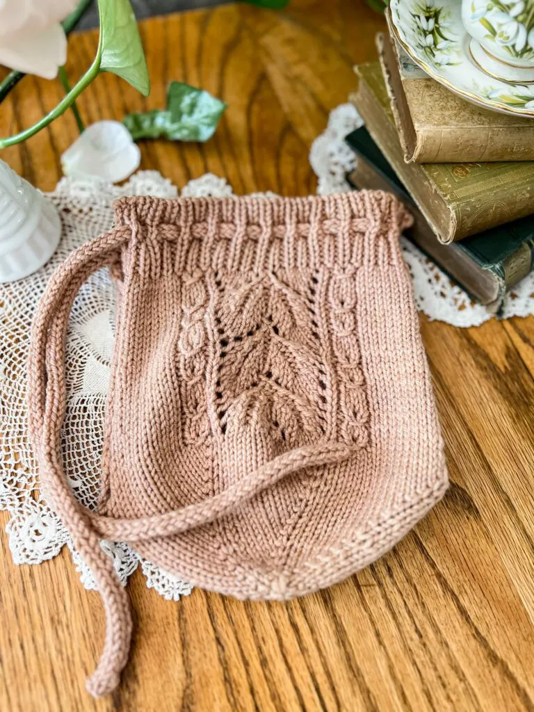 The Emily's Garden Tea Cozy is laid flat on a wooden table. This view shows how the bottom of the cozy is knit in a circle from the center outward before dividing into two halves for the sides of the cozy.