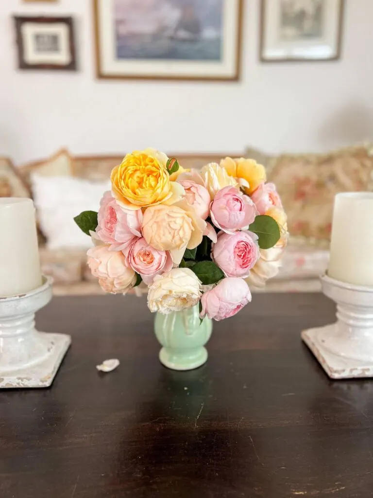 A light green vase full of pink, apricot, and yellow roses sits on a dark table flanked by two large candles.