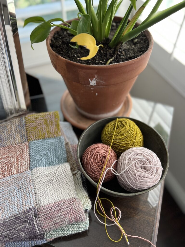 A slightly angled top-down image showing a metal bowl full of lavender, pink, and green balls of yarn sitting next to a scrappy blanket in progress. They're on a table in a sunny corner with a ceramic planter in the background.