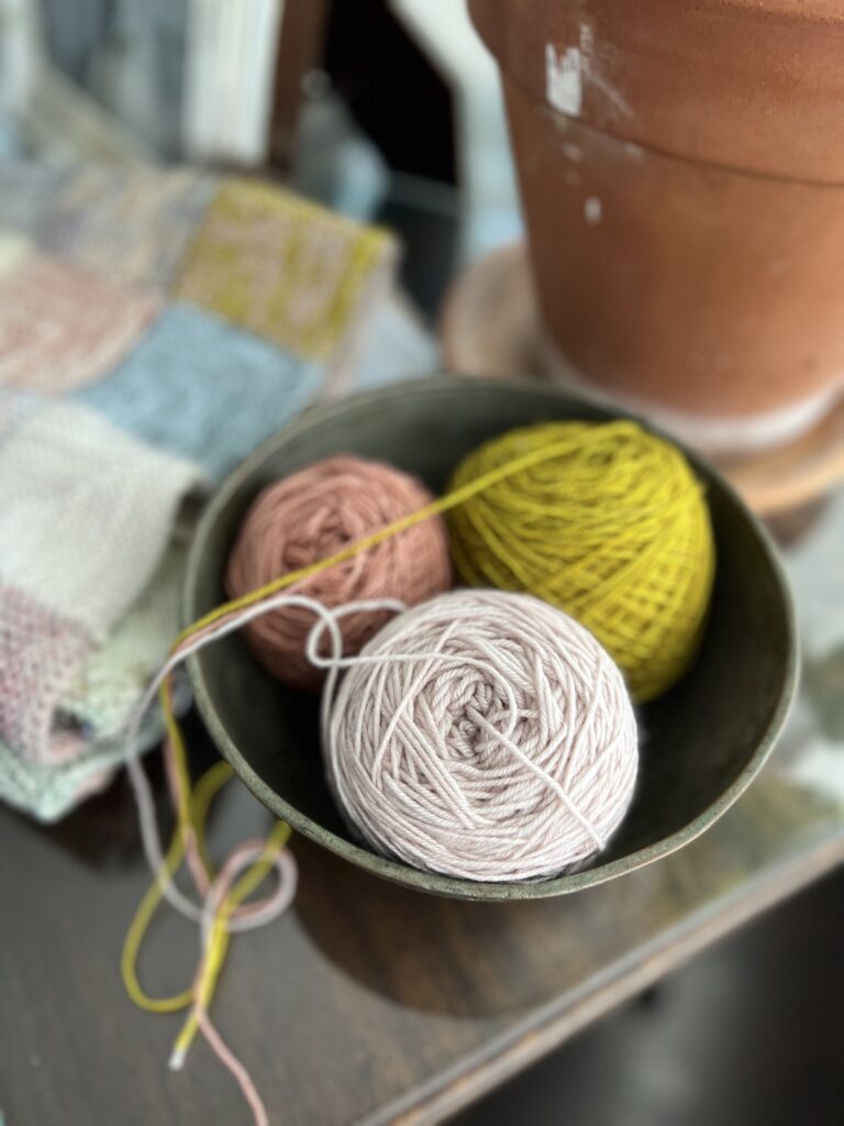 A top-down image of a metal bowl full of lavender, pink, and green yarn balls. Blurred in the background are a scrappy blanket in progress and a ceramic planter.