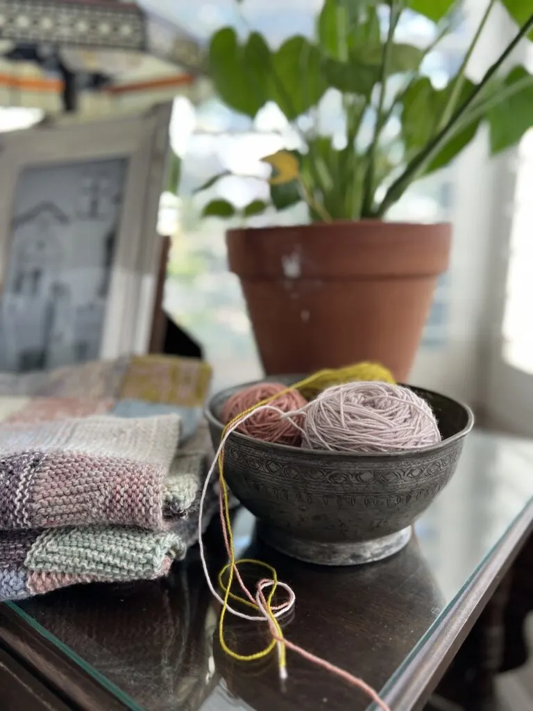 A sideways view of a metal bowl full of balls of yarn. In the background is a ceramic planter, and a scrappy blanket in progress sits to the side.