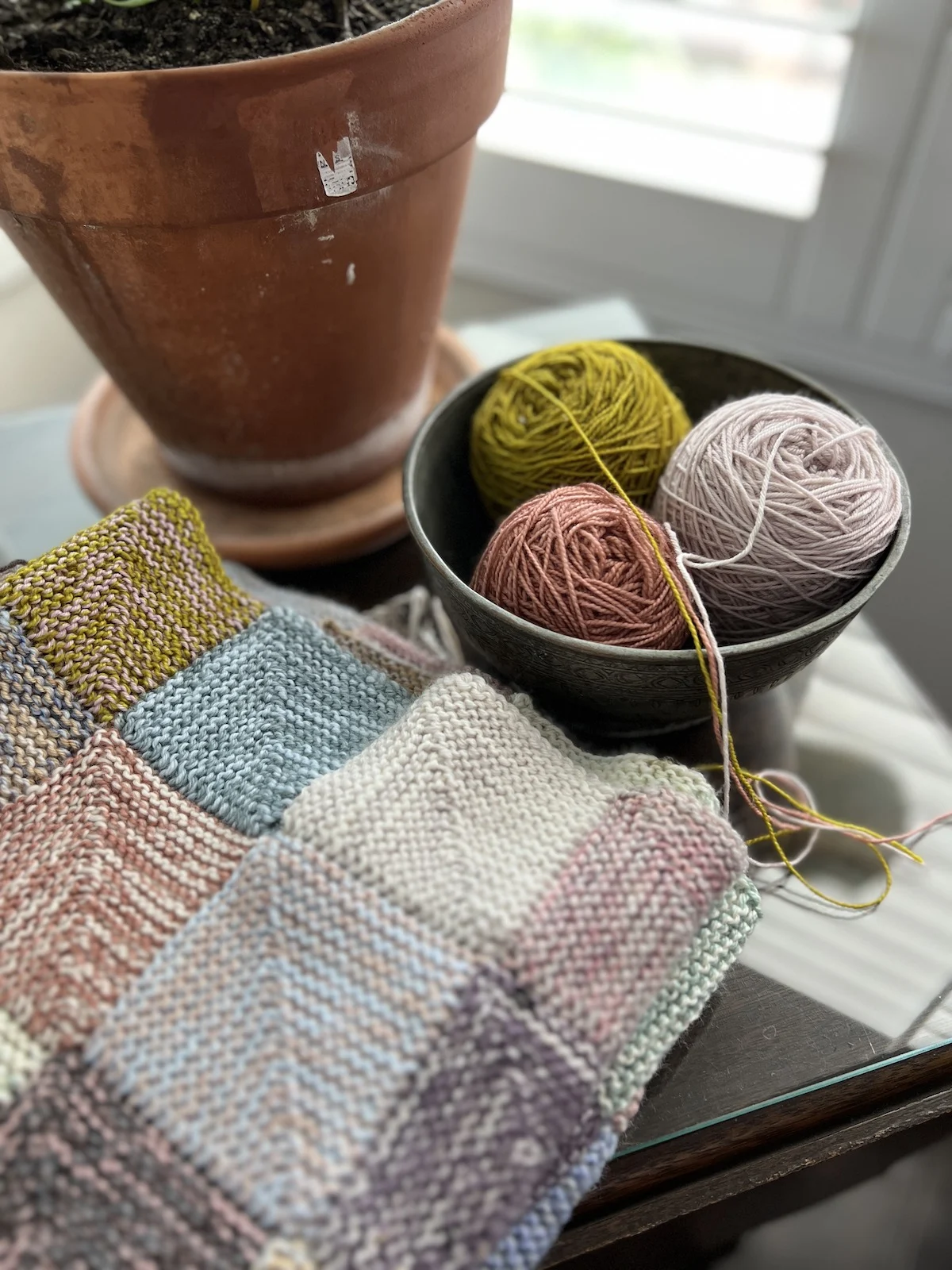 A metal bowl full of green, lavender, and pink yarn sits next to a ceramic planter. A multicolored mitered square blanket in progress sits in the foreground.