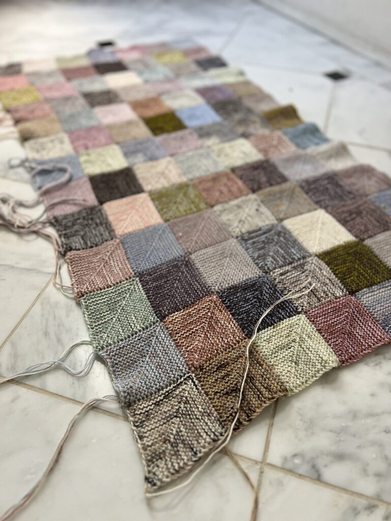 A close-up of the lower left corner of a mitered square blanket in progress. There are brown, blue, green, pink, purple, and tan squares.