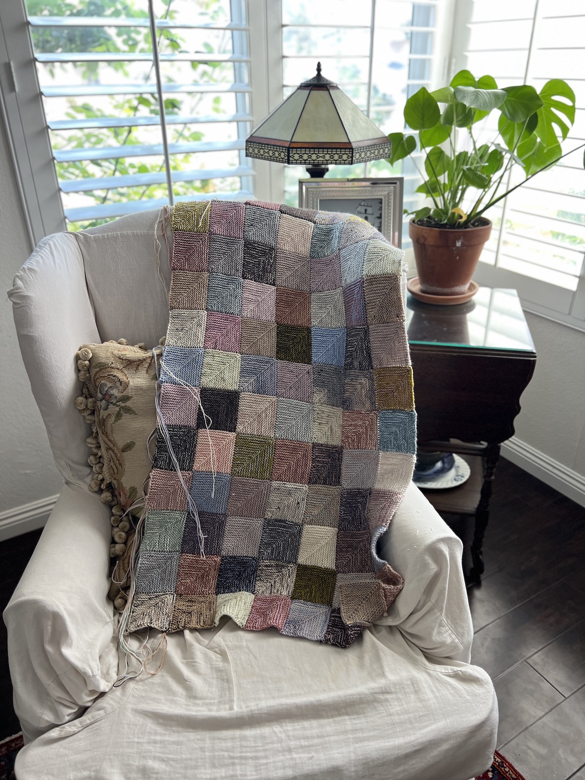 A multicolored mitered square blanket in progress is draped across the back of a slipcovered arm chair. In the background are a potted plant, a lamp, a picture frame, and some windows.