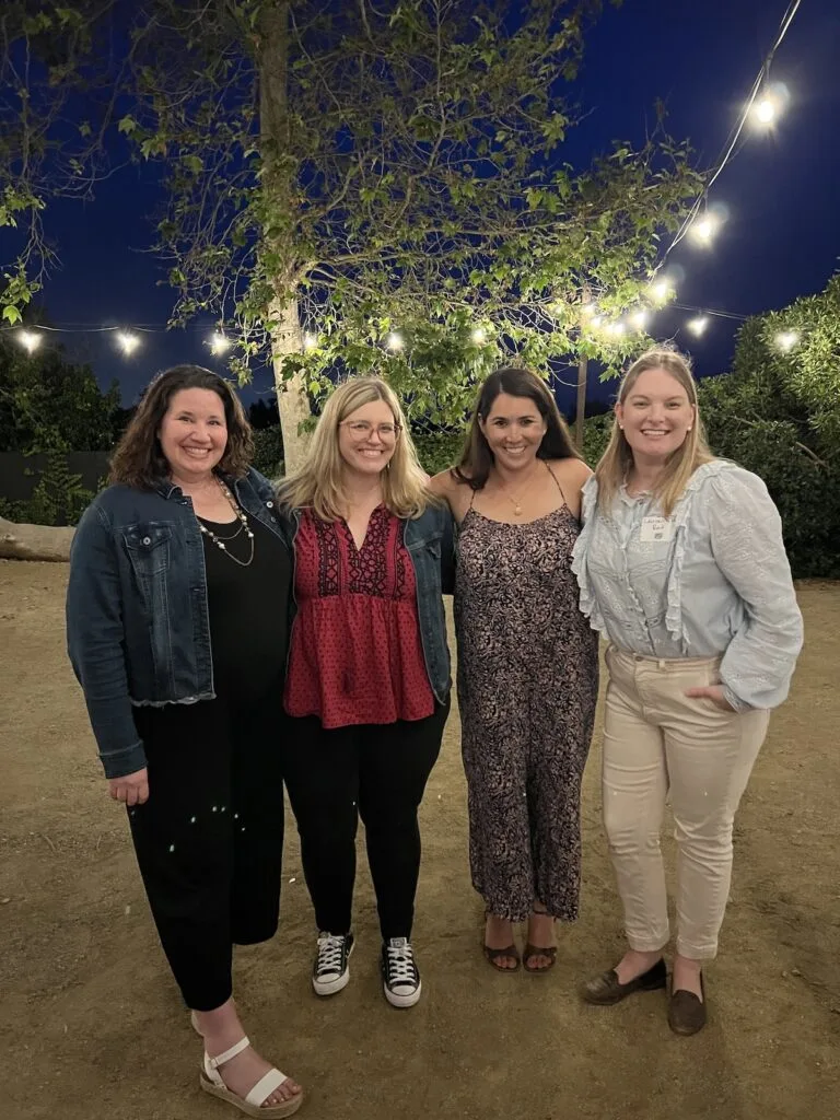 Four women in their late 30s stand with their arms around each other, grinning from ear to ear.