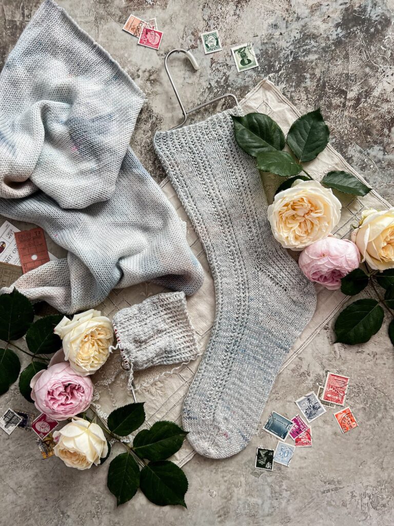 A top-down image of a finished sock knit from a sock blank, a sock in progress, the sock blank itself, some pink and cream roses, and some antique paper ephemera.
