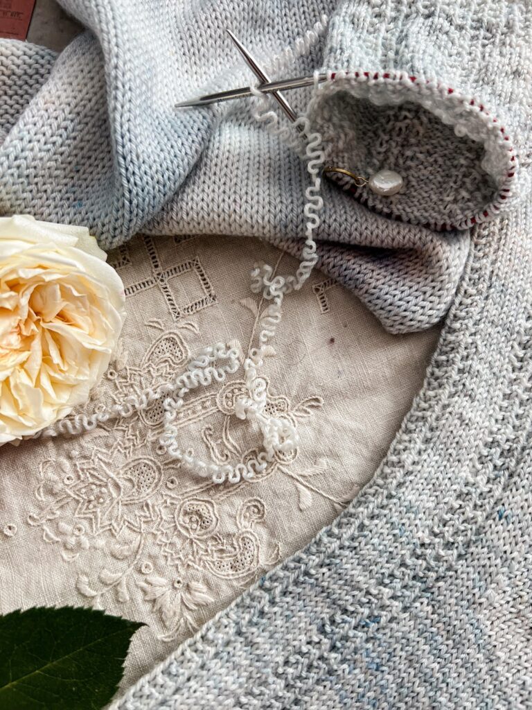 The crimped yarn from a sock blank is in focus in the center of the image, surrounded by one finished sock, a sock in progress, the sock blank itself, and a cream-colored rose.