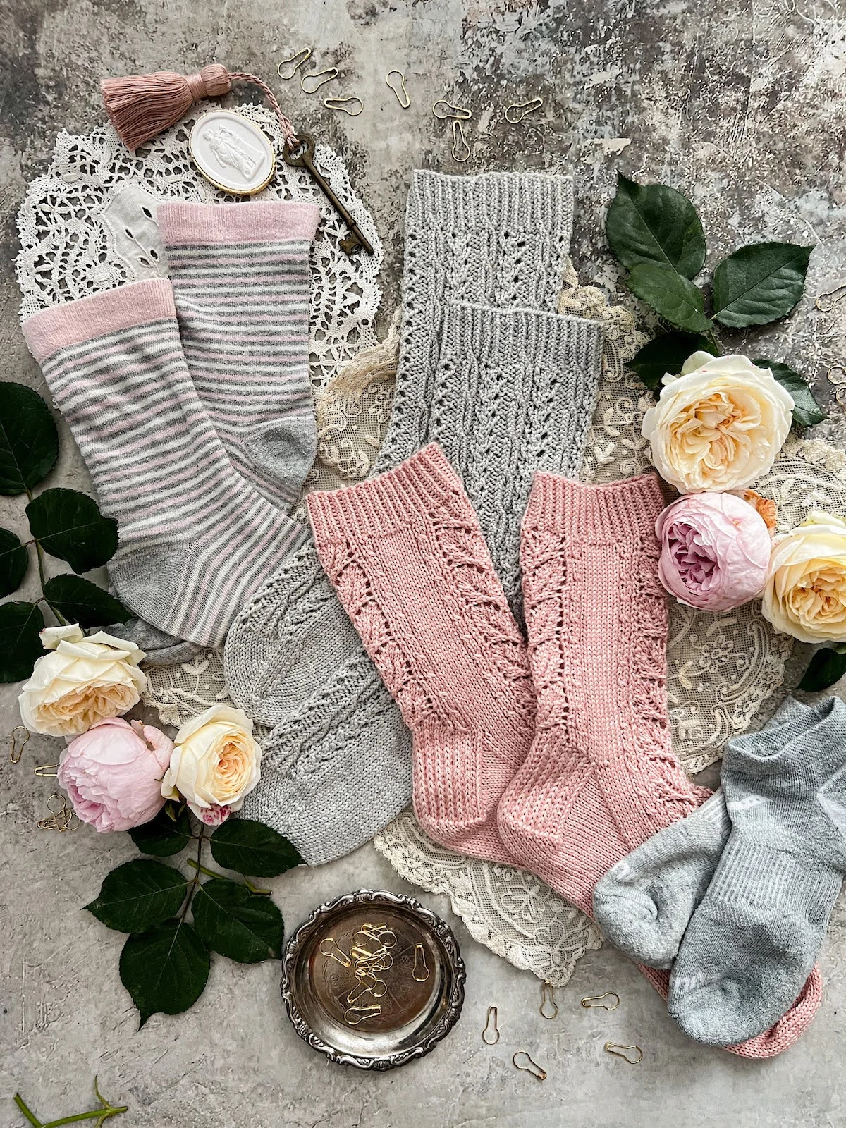 A top-down photo of two pairs of handknit socks (pink and gray) and two pairs of store-bought socks (gray athletic socks and mid-calf socks with stripes of pink, white, and gray). They're surrounded by pink and yellow roses, a plaster intaglio, a brass key with a pink tassel, and a tiny silver tray with brass bulb pins in it.