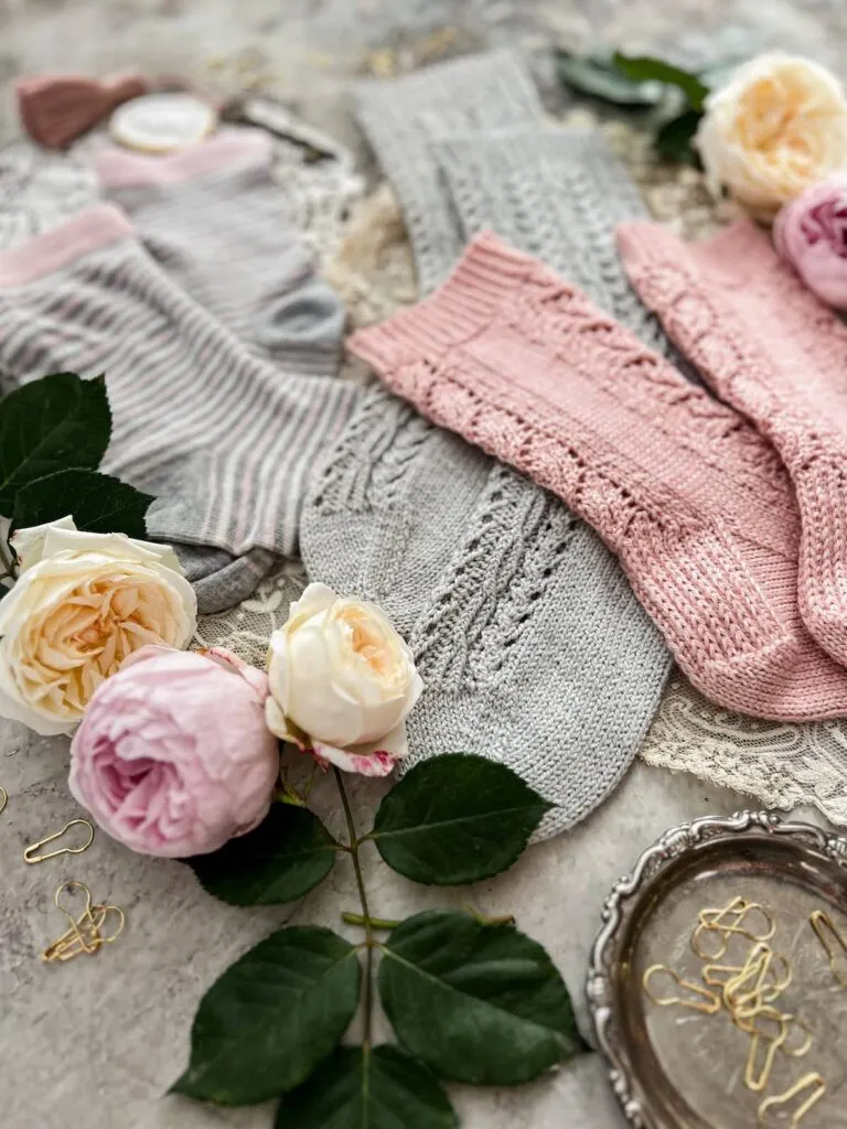 A close-up on the toes and heels of some hand-knit and store-bought socks in shades of pink and gray. There are pink and yellow roses in the foreground.