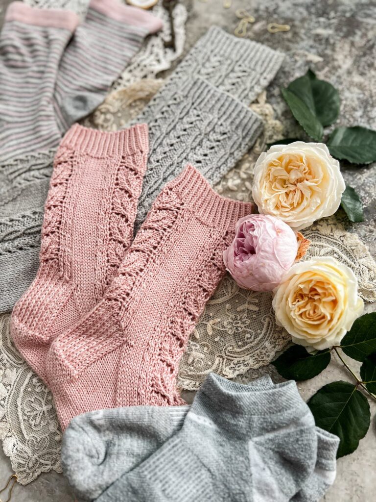 A pair of pink hand-knit socks is in focus in the center of the image, along with some pink and yellow roses. They're surrounded by gray handknit and store-bought socks that are blurred at the edges of the image.