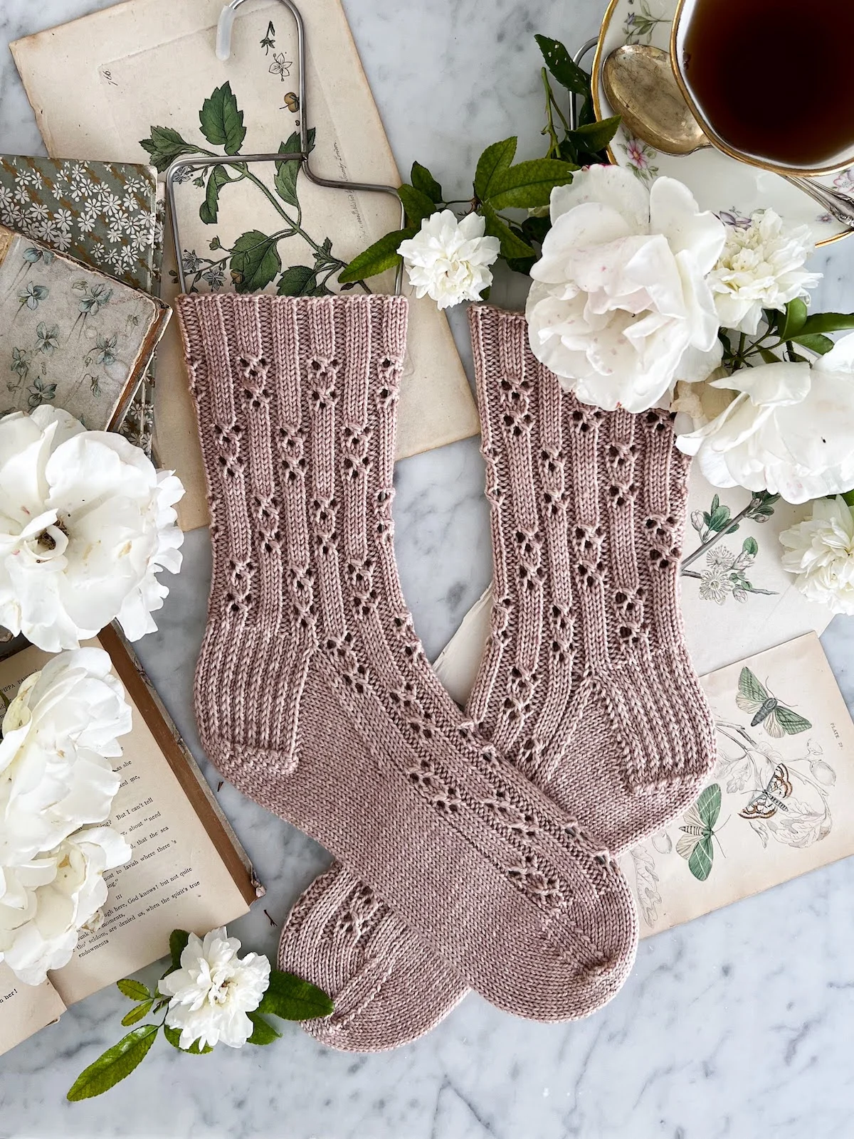 A top-down photo of a pair of pink, handknit socks laid flat on a white marble countertop. The socks are crossed at the foot and feature an all-over eyelet pattern. They're surrounded by antique paper ephemera, white roses, old books, and a teacup full of tea.