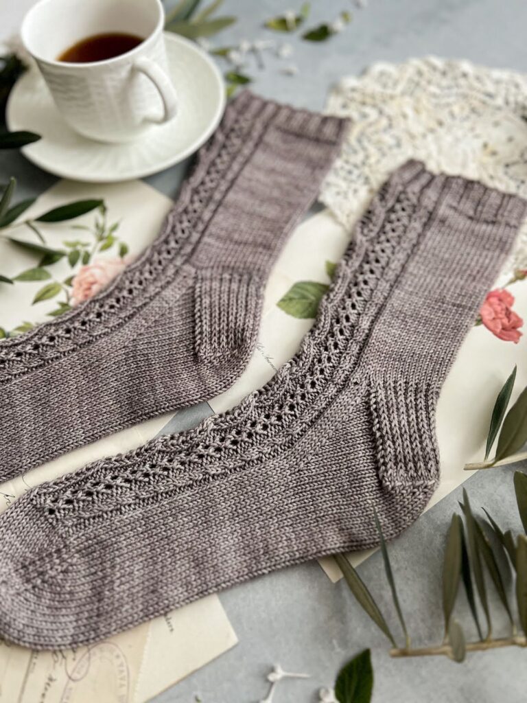 A close-up on the foot of a grayish-purple sock with cables and eyelets down the front. Its mate is in the background. They're surrounded by antique paper ephemera and a white teacup full of espresso.