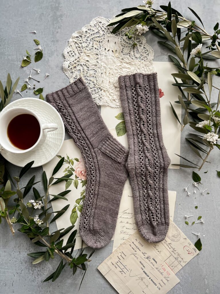 A pair of grayish-purple socks is laid out flat on a gray surface. One sock is turned to the left, and the other is laid out so the front panel is visible all the way from cuff to toe. They have cables and eyelets running down the front of each sock. The socks are surrounded by antique paper ephemera, textiles, olive branches, and a white teacup full of espresso.