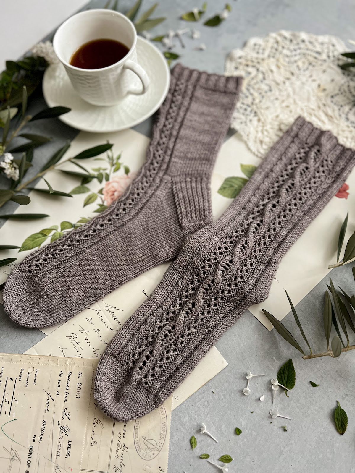 A pair of grayish-purple socks is laid out flat on a gray photo background, surrounded by antique paper ephemera and a white teacup full of espresso. This photo is shot slightly from the left and focuses on the sock that clearly shows the textured panel all the way down from cuff to toe.