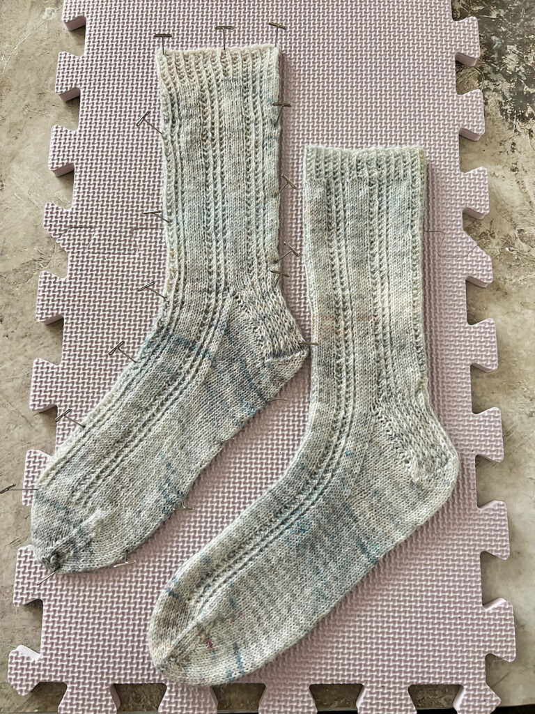 Two light blue socks blocking on a light purple blocking mat. One sock is just laid flat, while the other has been pinned into place.