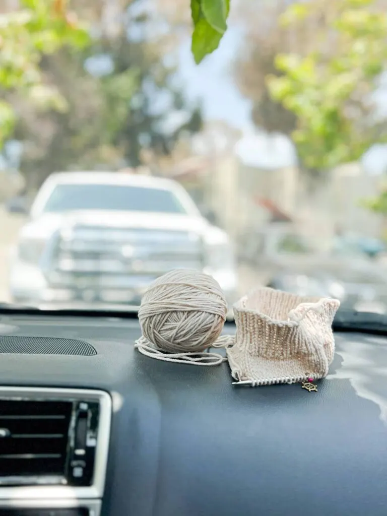 A ball of tan yarn and a half-knit hat sit on the black dashboard of a car. A parked truck is blurred in the background.
