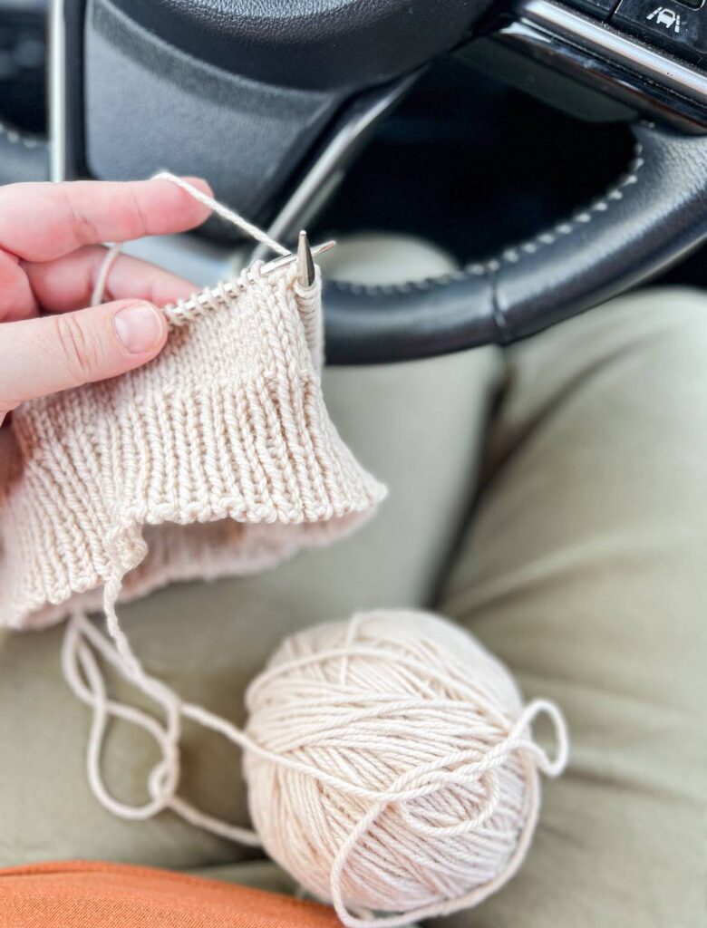 A ball of tan yarn and a half knit hat in the lap of a driver, with the steering wheel just visible in the top corner. Don't knit while actually driving! This photo was taken while parked in a parking lot.