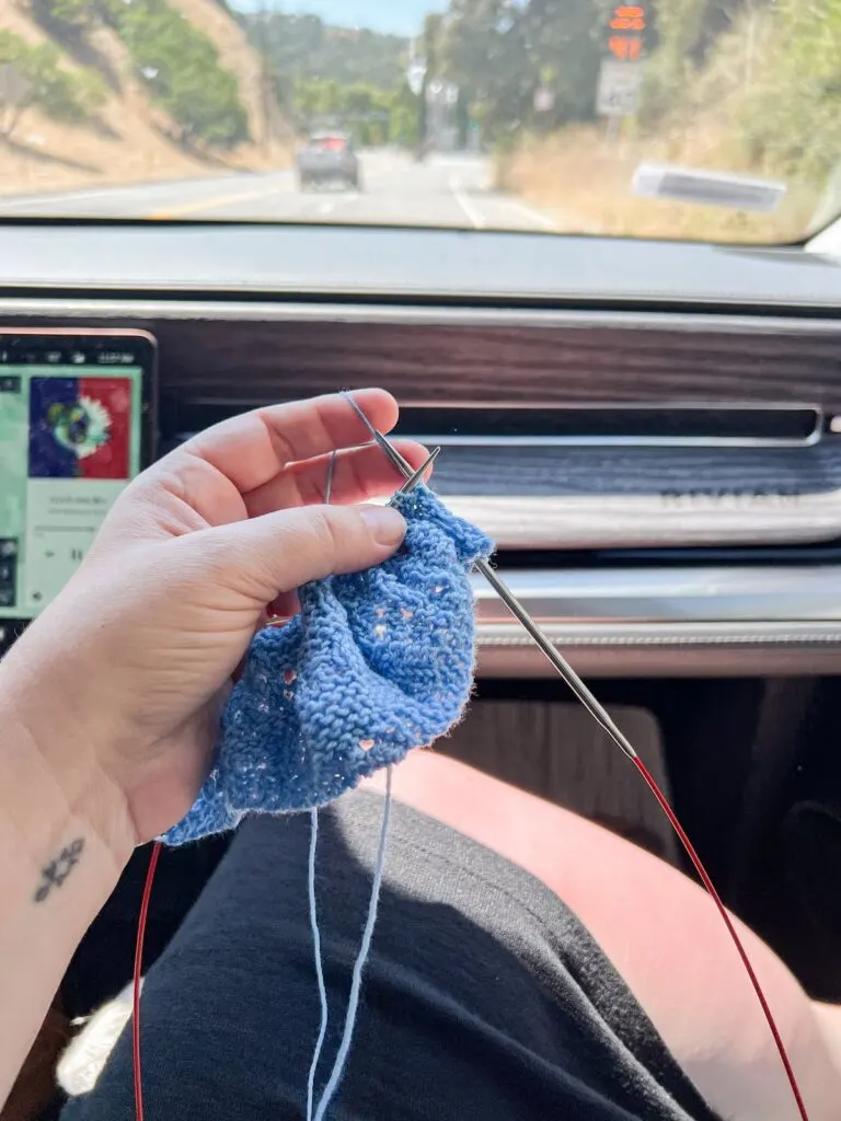 Holding up some light blue knitting with my left hand while riding in the passenger's seat of my friend's car. Through the windshield you can see the sun-browned California summer landscape.
