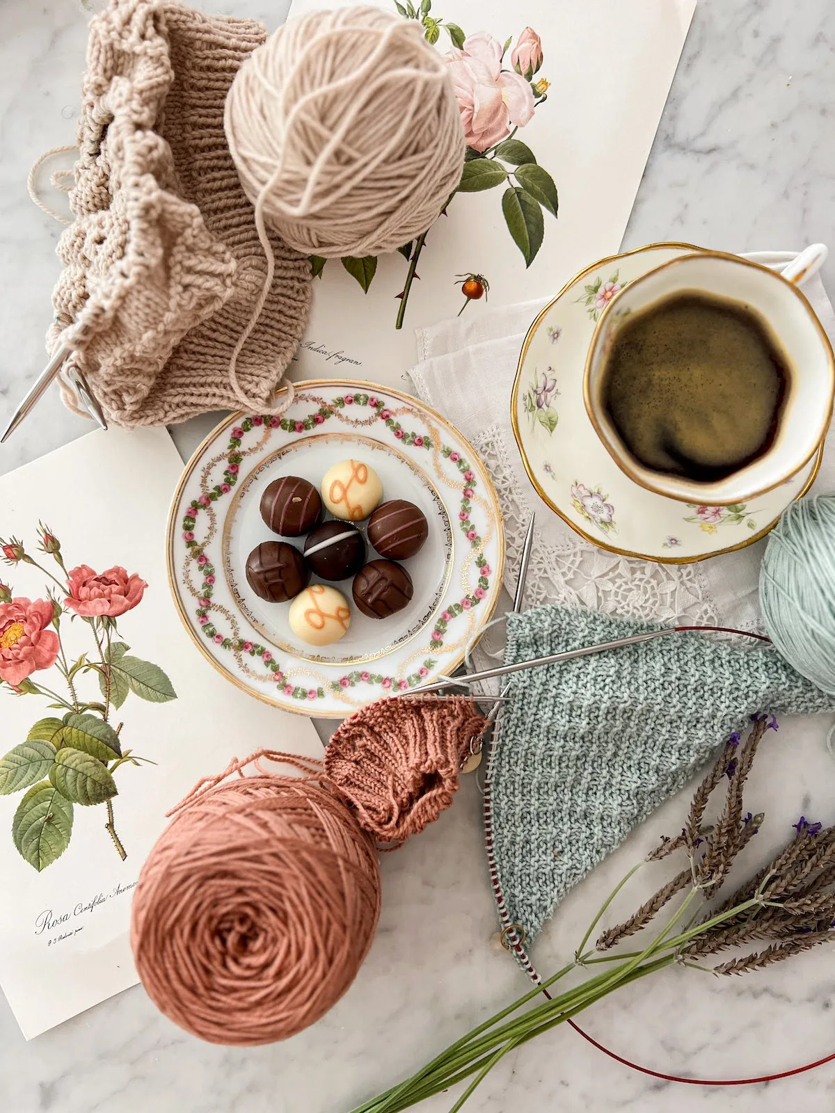 A top-down photo of three knitting projects in progress, all on circular needles. They are a tan hat, a dark pink sock, and a mint shawl. They're surrounded by vintage botanical prints, an ornate plate with chocolate bonbonbs on it, and a pastel teacup full of espresso.