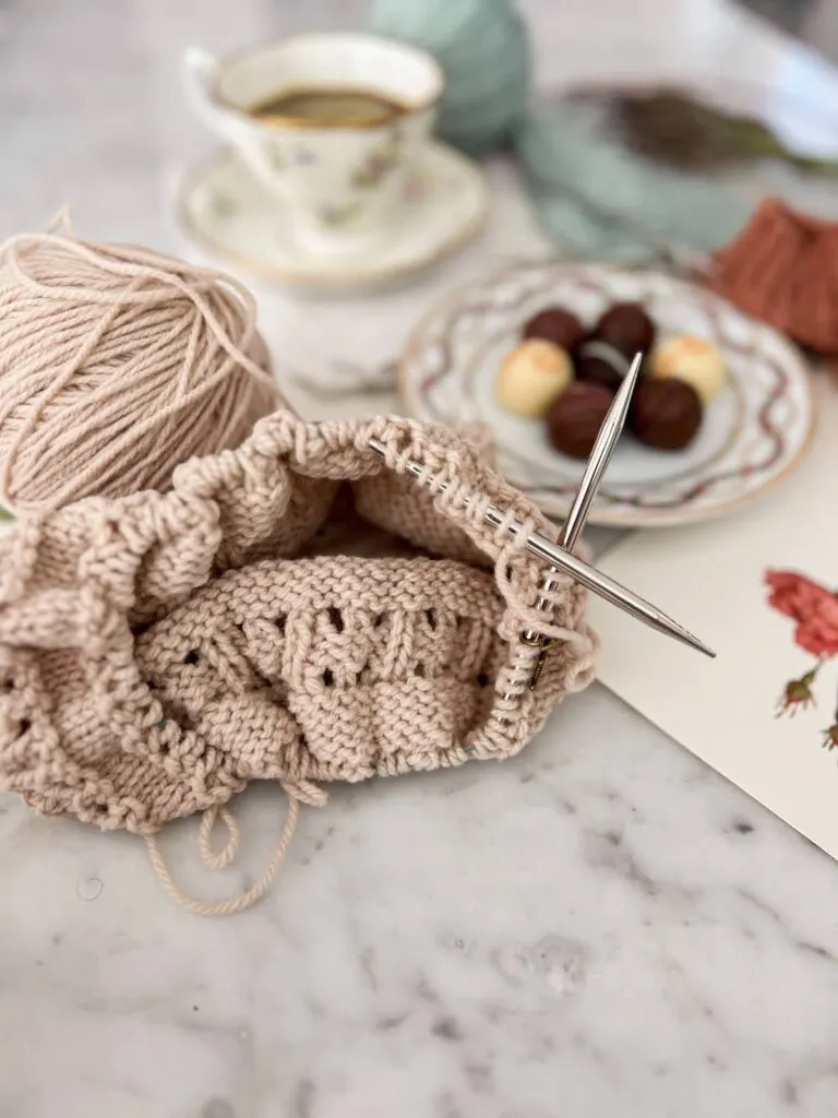 A close-up of a tan hat in progress. It's being knit on circular needles. The needles are pointing outward to the right.