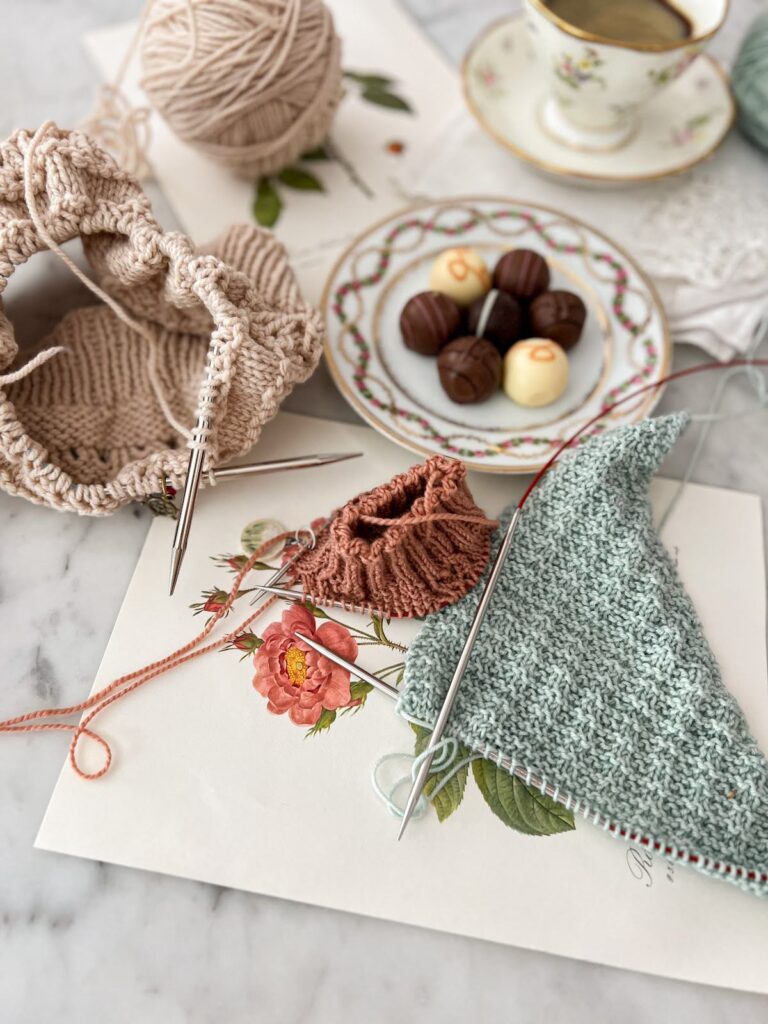 A side-angled photo showing three knit projects (a tan hat, a pink sock, and a mint shawl) all knit on circular needles. In the background are vintage botanical prints, an ornate plate with bonbons on it, and a pastel floral teacup full of espresso.