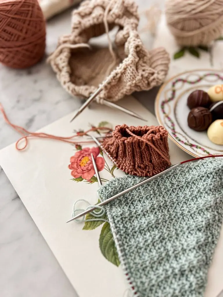 Three projects on circular needles are lined up from top to bottom. At top, there is a tan hat in progress. In the middle is a dark pink sock on tiny circular needles. At the bottom is a mint shawl on very long circular needles. They are on top of a vintage botanical print and next to an ornate little plate full of chocolates.
