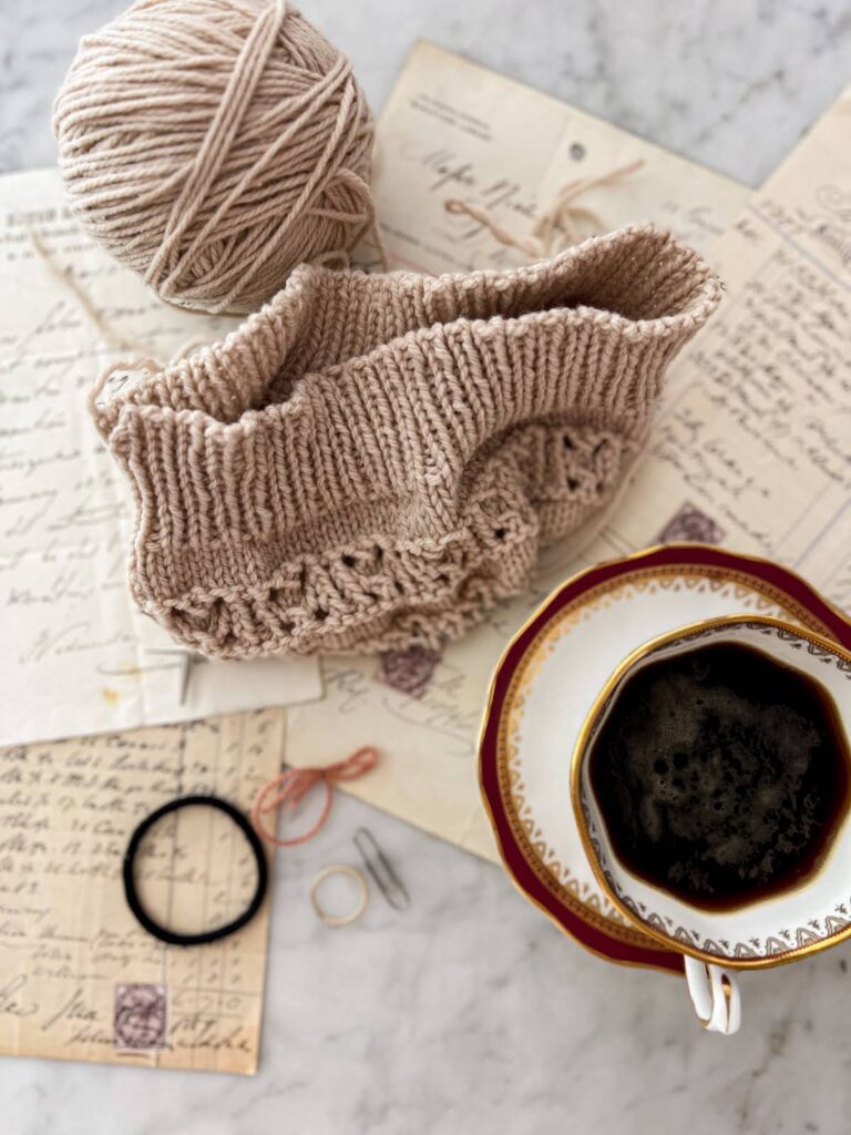A top-down photo of a half knit hat in progress, a teacup full of espresso, some antique paper ephemera, and a few bits and bobs that can serve as stitch markers in a pinch: a hair tie, a ring from my right hand, a paper clip, and a loop of yarn tied off.