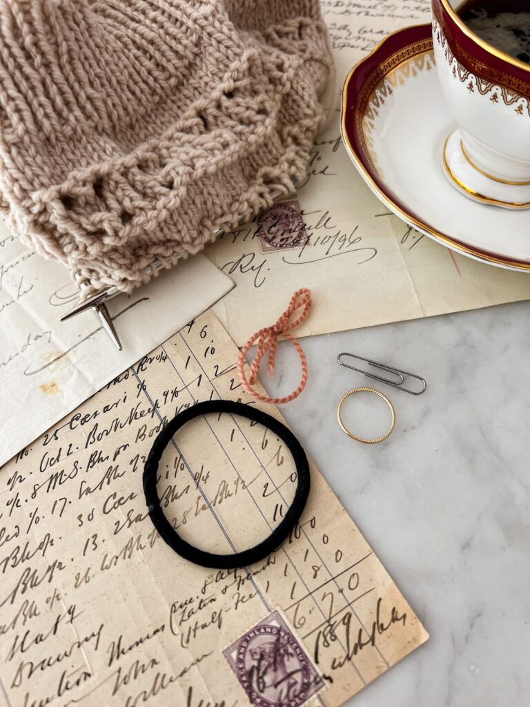 A close-up look at what could be junk at first glance, but is in fact a small collection of items that can work as stitch markers in a pinch: a hair tie, a ring from my right hand, a paper clip, and a loop of yarn tied off. They're surrounded by antique paper ephemera, a tea cup, and a tan hat in progress.