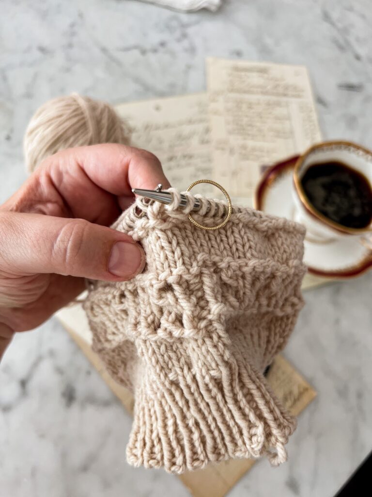 A close-up on my left hand holding a tan hat in progress. A gold ring has been placed onto the needle to serve as a stitch marker. Blurred in the background are some antique paper ephemera and a teacup.