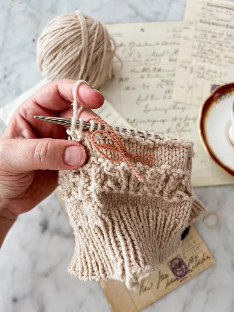 A close-up on my left hand holding a tan hat in progress. A loop of pink yarn is tied onto the needle to serve as a stitch marker. Blurred in the background are some antique paper ephemera and a teacup.