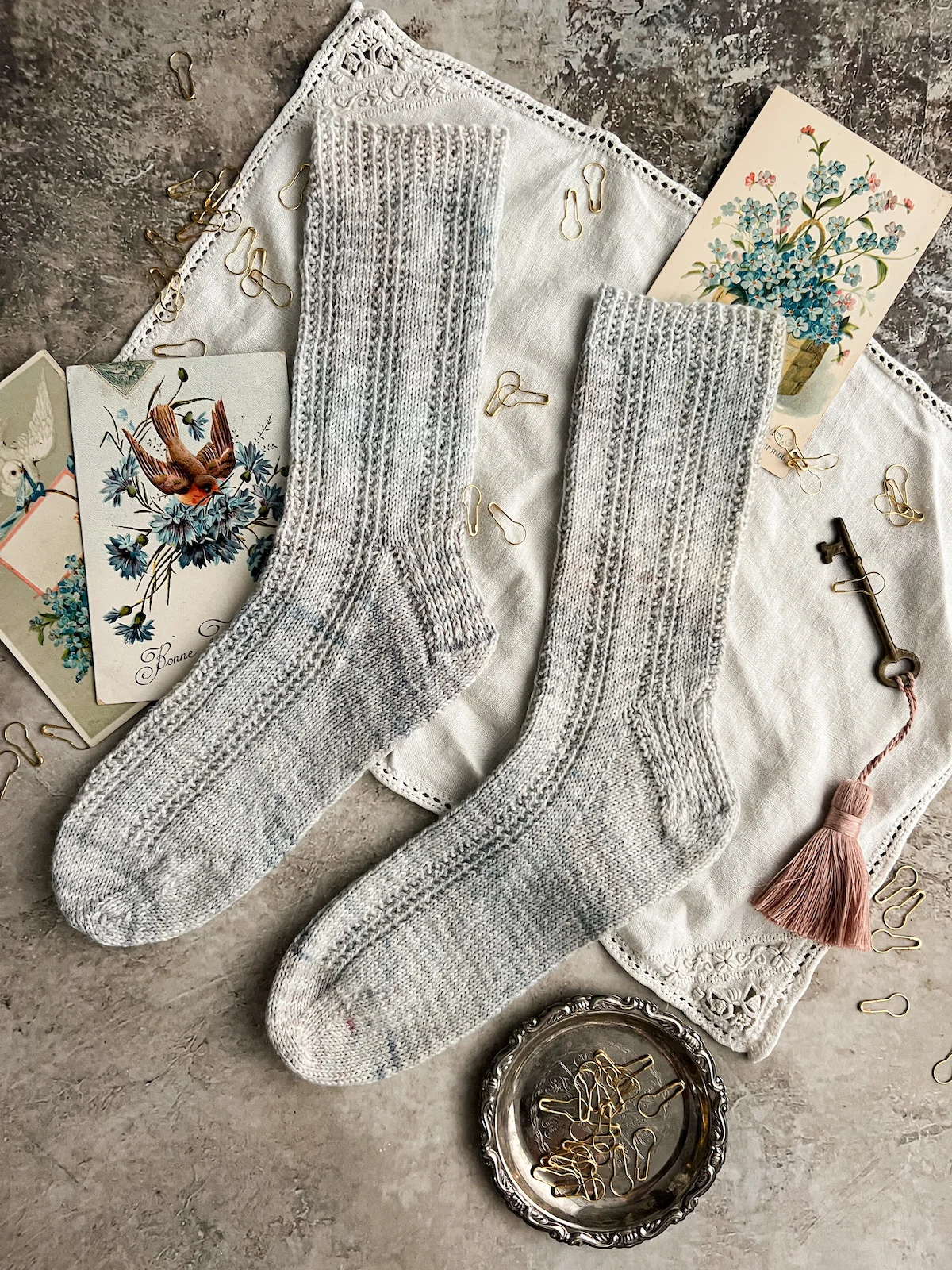 A top-down photo of a pair of light blue socks laid flat on an antique lace-trimmed placemat. The socks have columns of purls running up and down the legs and are pointing to the left. They are surrounded by antique postcards with blue flowers on them, a brass key with a pale pink tassel, and a silver tray full of brass bulb pins.
