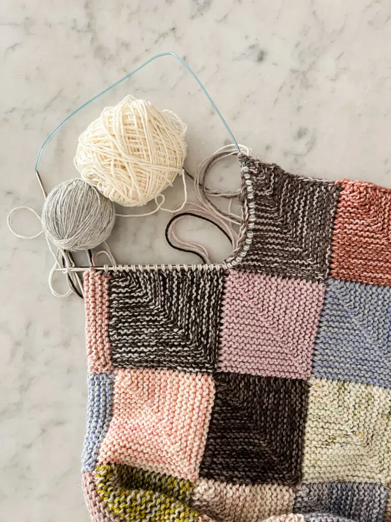 A top-down photo of a multi-colored scrappy mitered square blanket, showing how the stitches have been picked up along the edges of two squares to start knitting a third square.