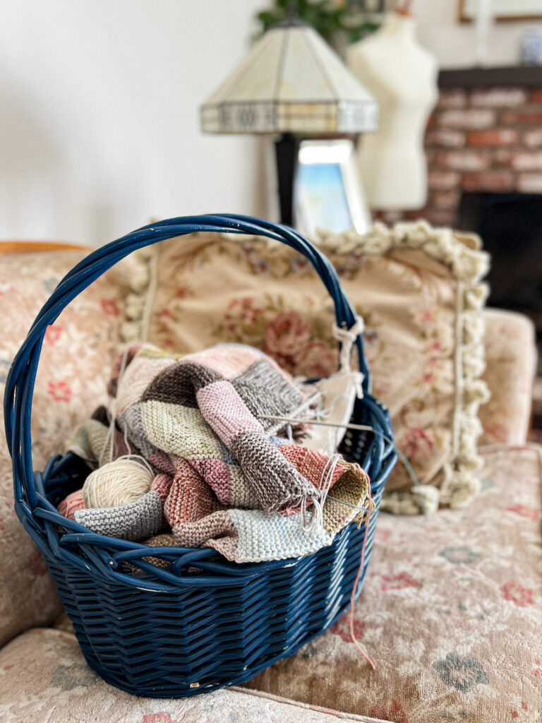 A dark blue wicker basket sits on an antique floral couch. The basket is filled with scrap balls of yarn and a multi-colored mitered square blanket in progress.