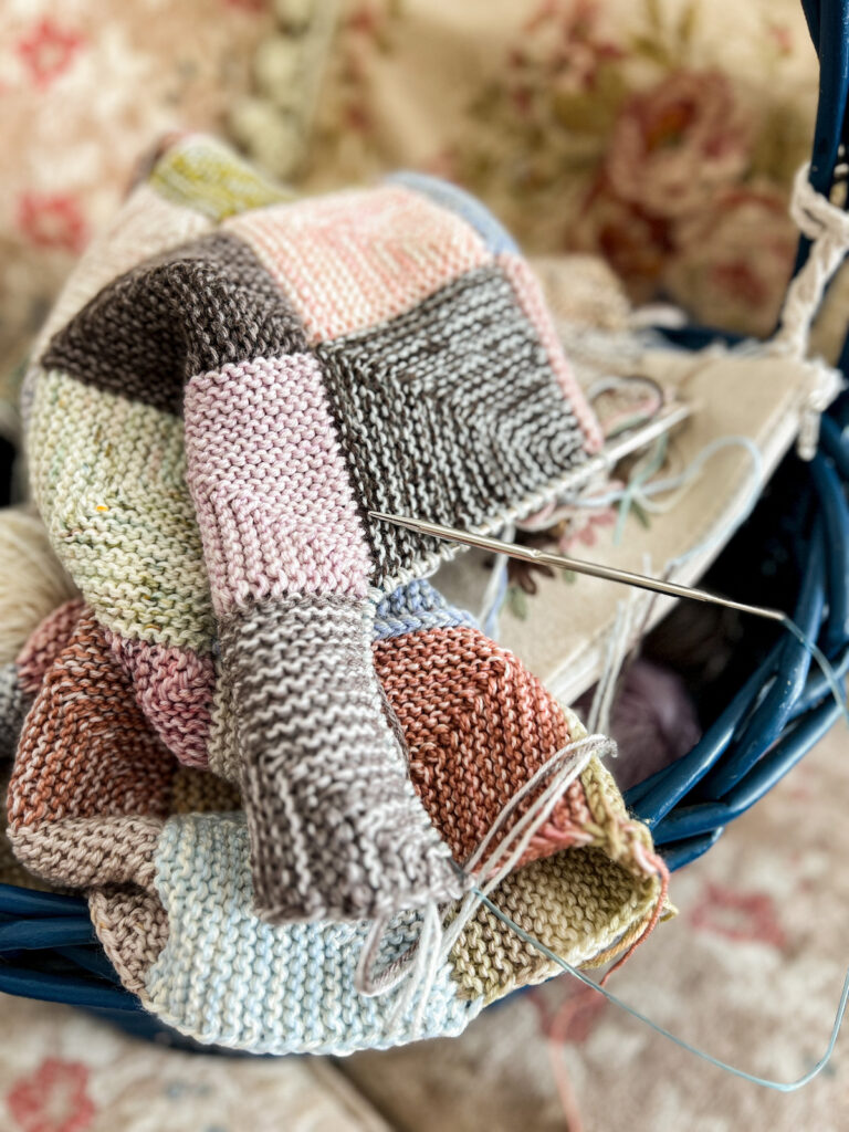 A close-up photo of a multi-colored mitered square blanket in progress. The blanket is sitting in a dark blue wicker basket.