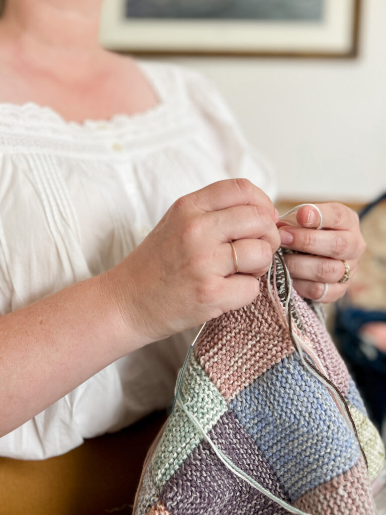 A white woman (me!) wearing a white poplin blouse with pintucks and lace trim works on knitting a multi-colored scrappy mitered square blanket. I'm knitting continental style, with the working yarn tensioned around my left hand.