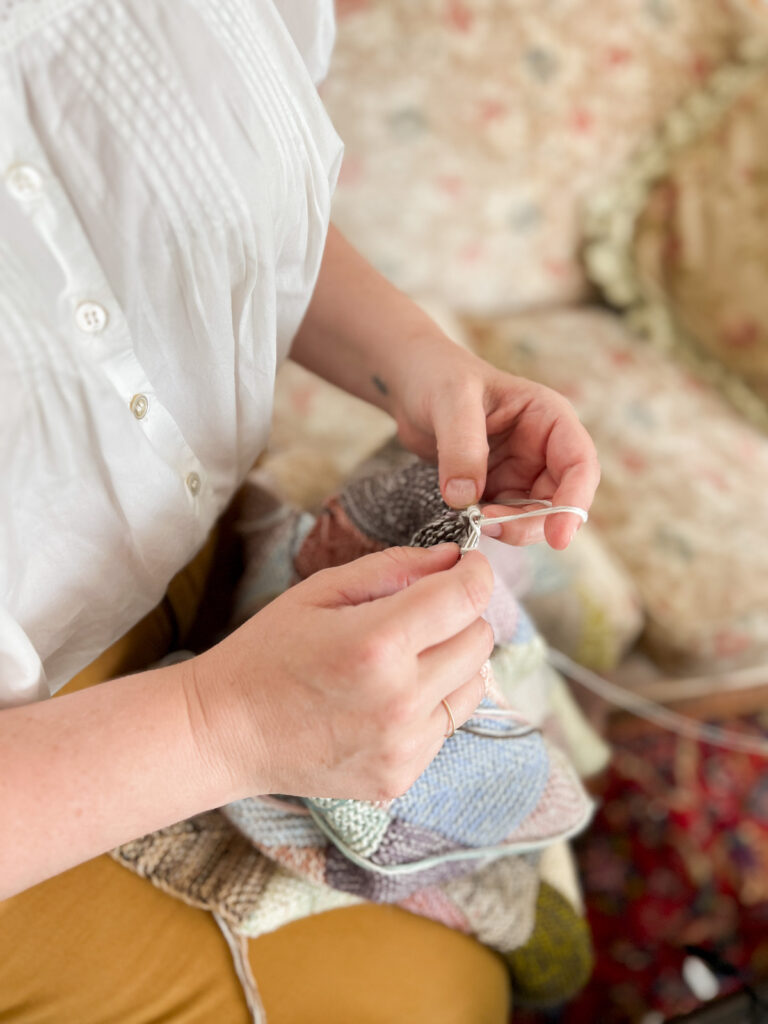 A top-down image of a white woman's hands (mine!) as she works on knitting a multi-colored mitered square blanket. I'm tensioning the yarn around my left hand and sitting on my great-grandmother's floral sofa.