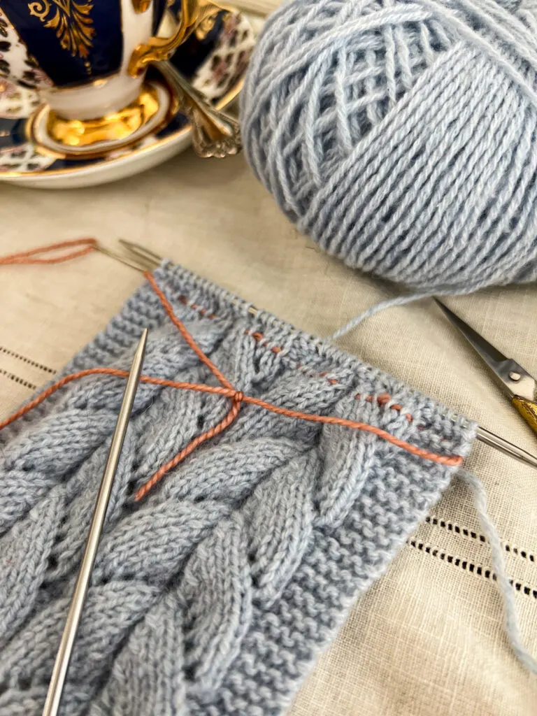 A close-up on a piece of light blue lace knitting, showing how the pink lifeline threaded through at the top of the work is slightly visible.