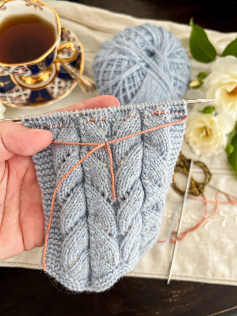 A white woman's left hand (mine!) holds a piece of light blue knitting in the forefront of the picture. The picture focuses on the pink lifeline threaded through the stop stitches and shows how the lifeline has been tied in front of the work to secure it.