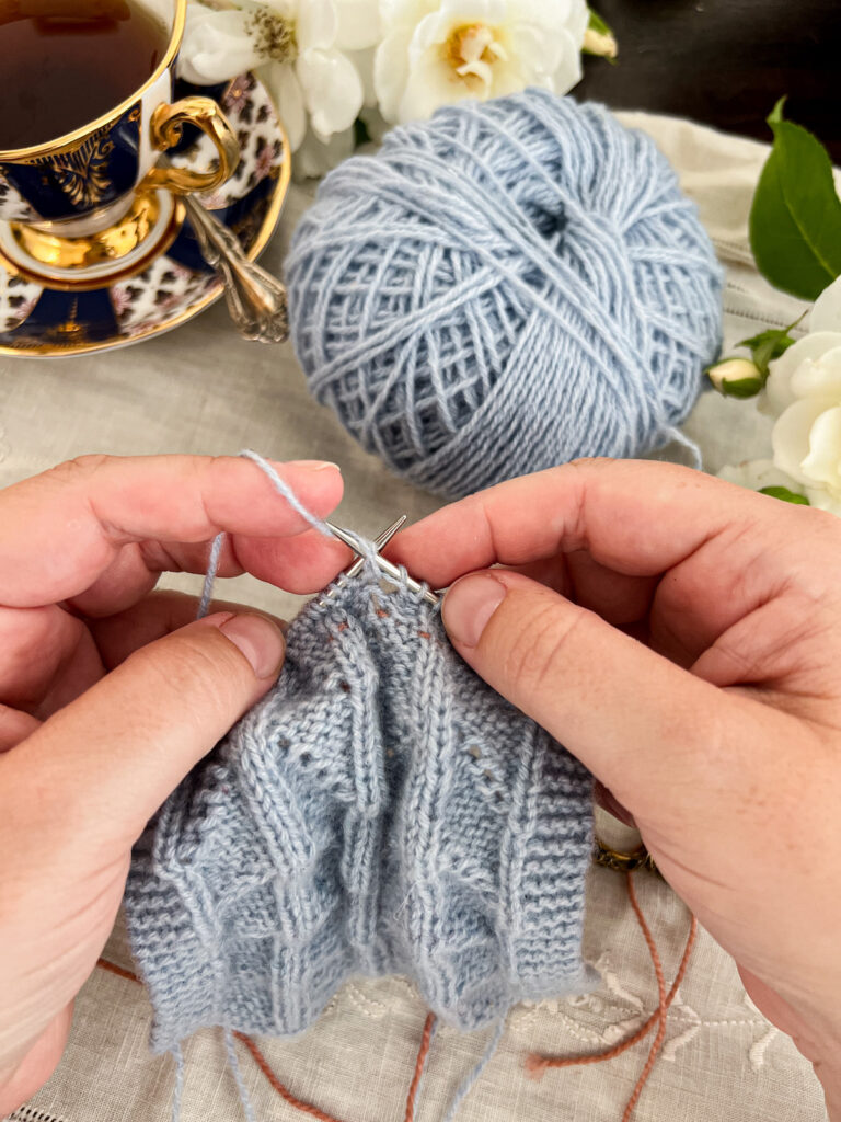 A white woman's hands (mine!) knit on the reverse side of a piece of light blue lace knitting. This photo shows how the pink lifeline is almost invisible from the back side.