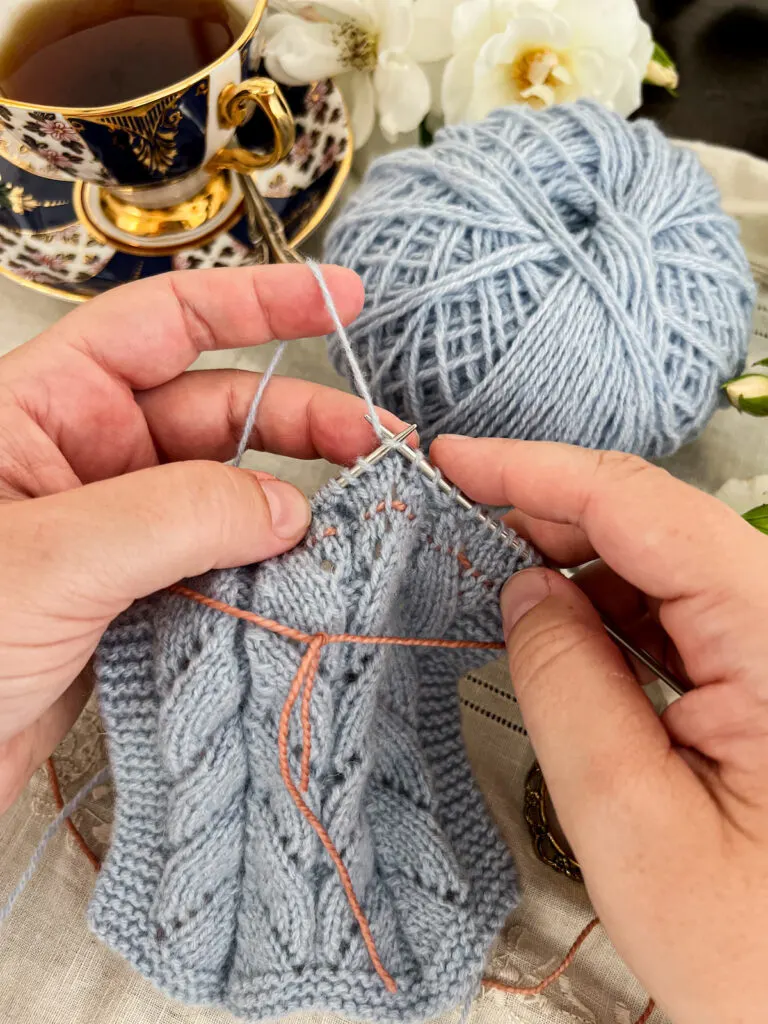 A white woman's hands (mine!) knit on the front side of a piece of light blue lace knitting. The photo focuses in on how the lifeline is more visible when working on the front side of the yarn.