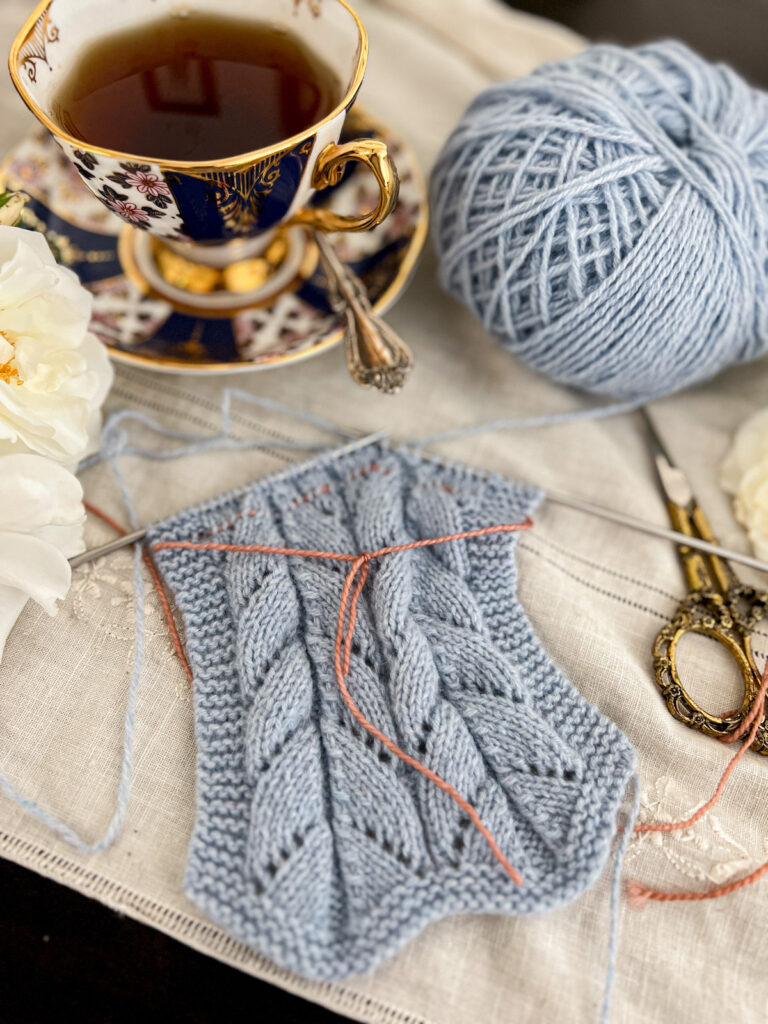 A top-down image of a piece of light blue lace knitting in progress, with the row half-knit. A pink lifeline has been threaded through a row of stitches near the top, but a few rows have been worked since the lifeline was placed. The knitting is surrounded by ornate brass scissors, a ball of light blue yarn, a blue and gold teacup full of tea, and some white roses.