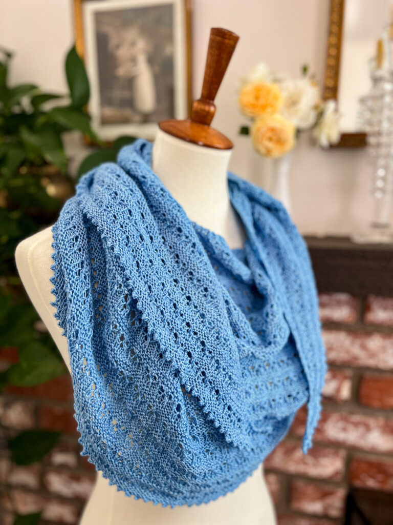 A side-view of a light blue, crescent-shaped shawl draped around a dressmaker's form. The shawl features lots of eyelets and garter stitch for abundant texture.