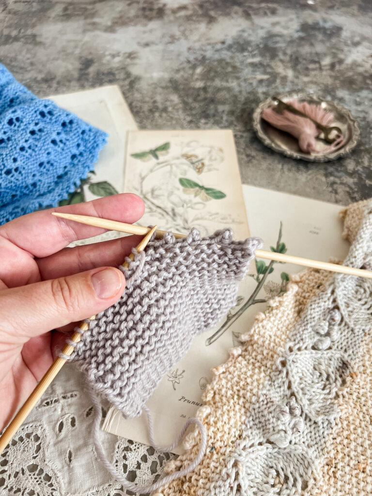 A white woman's left hand (mine!) holds a gray garter-stitch square that is half bound-off. There are four picots to the right and all the other stitches are still on the working needle.